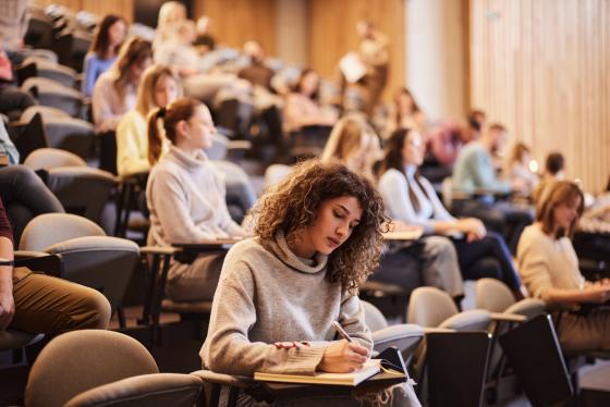 En kvinnelig student sitter i et auditorium og skriver med mange medstudenter bak seg