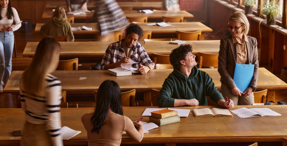 Blid, kvinnelig professor snakker med mannlig student i biblioteket med flere studenter rundt